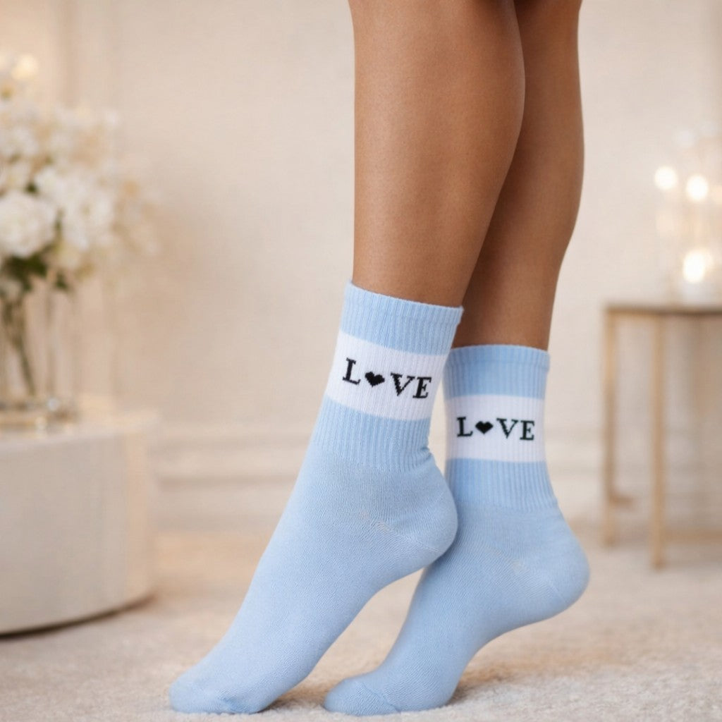 Hellblaue Socken mit weißem Streifen und schwarzem „LOVE“-Schriftzug (Herz statt O) an den Füßen einer stehenden Person, aufgenommen in einem hellen, wohnlichen Innenraum mit weichem Hintergrund.