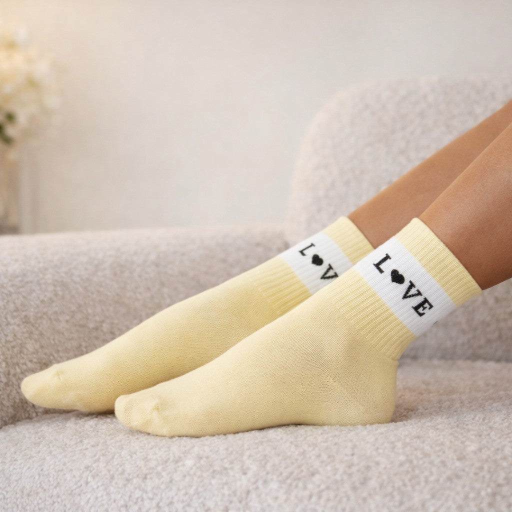 Hellgelbe Socken mit weißem Bündchen und schwarzem „LOVE“-Schriftzug (Herz statt O) an den Füßen einer liegenden Person auf einem hellen, gepolsterten Sofa in weichem Innenlicht.