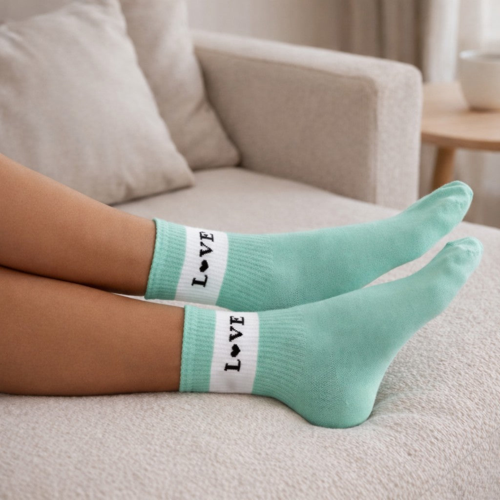 Mintgrüne Socken mit weißem Bündchen und schwarzem „LOVE“-Schriftzug (Herz statt O) an ausgestreckten Füßen einer liegenden Person auf einem hellen Sofa in wohnlicher Umgebung.