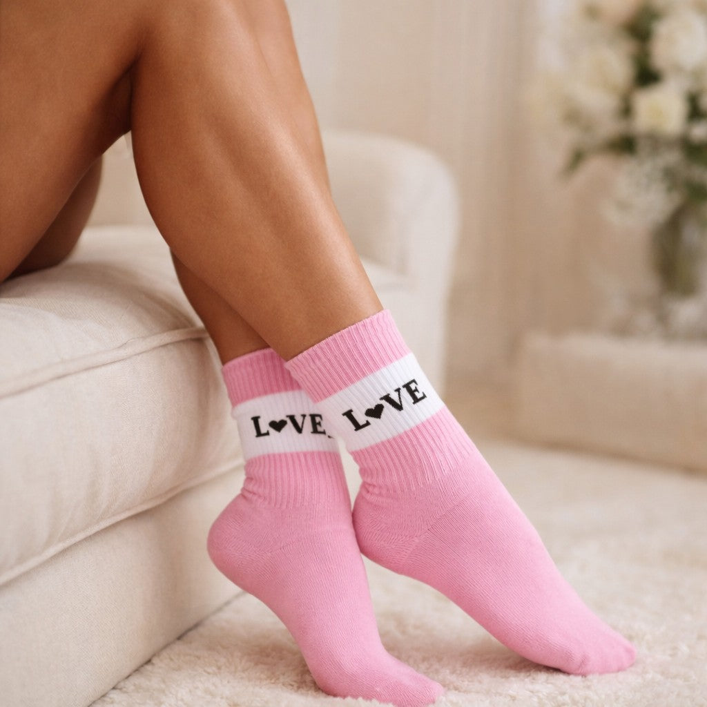 Rosa Socken mit weißem Bündchen und schwarzem „LOVE“-Schriftzug (Herz statt O) an leicht angewinkelten Füßen einer sitzenden Person auf einem hellen Sofa in wohnlicher Umgebung.